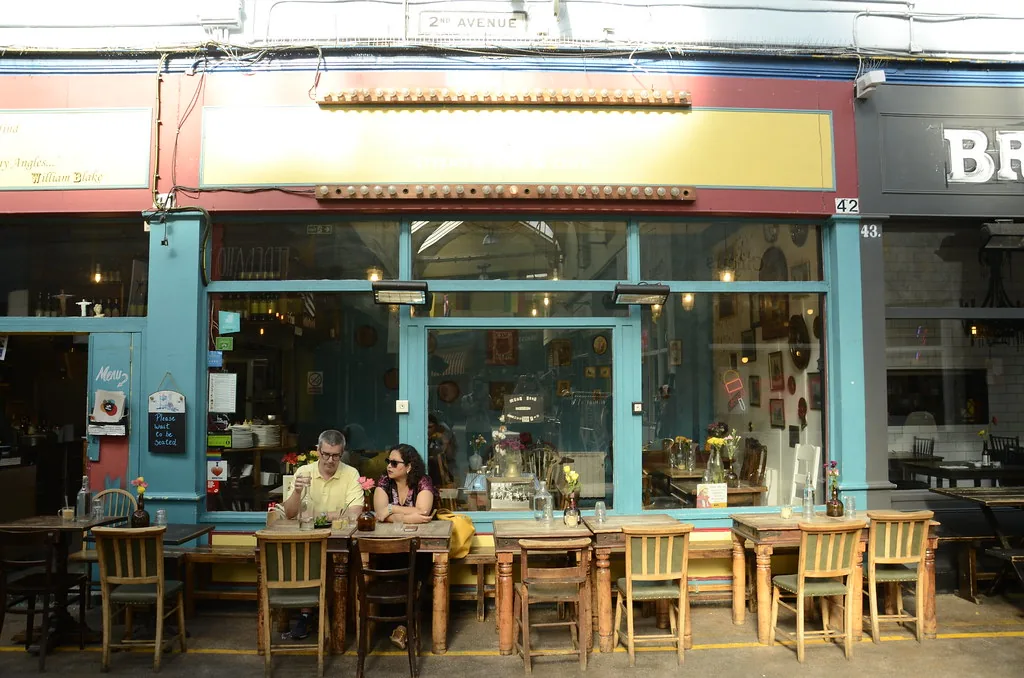 Diners at communal tables under the 2nd Avenue arcade sign in Brixton Village covered market