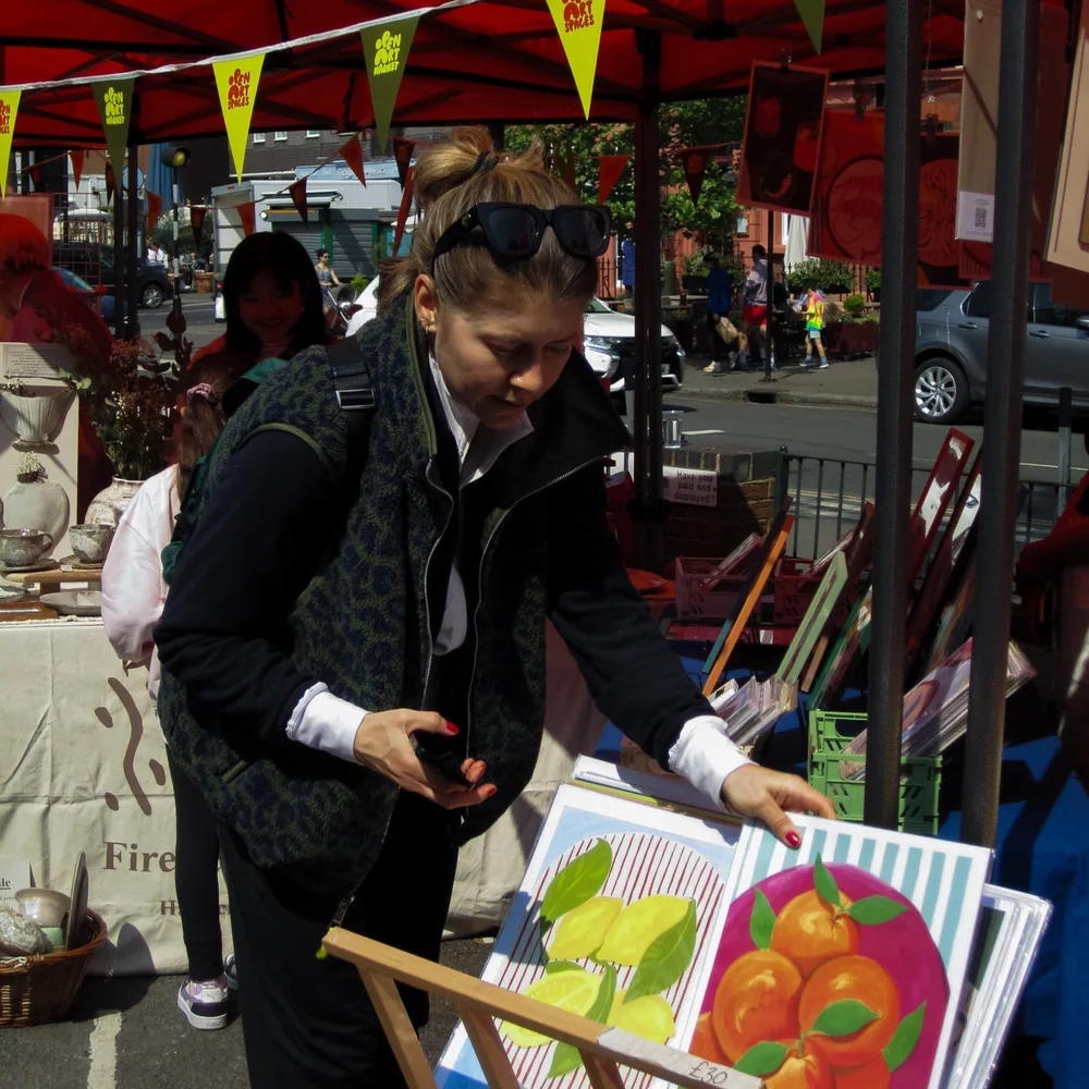 Shopper browsing framed original artwork and prints under an orange stall canopy with Open Art Spaces bunting at the Open Art Market on Portobello Road